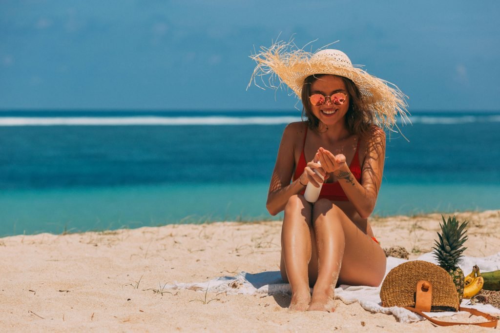 Woman in straw hat and sunglasses applying sunscreen on a sunny beach, enjoying a relaxing summer day.